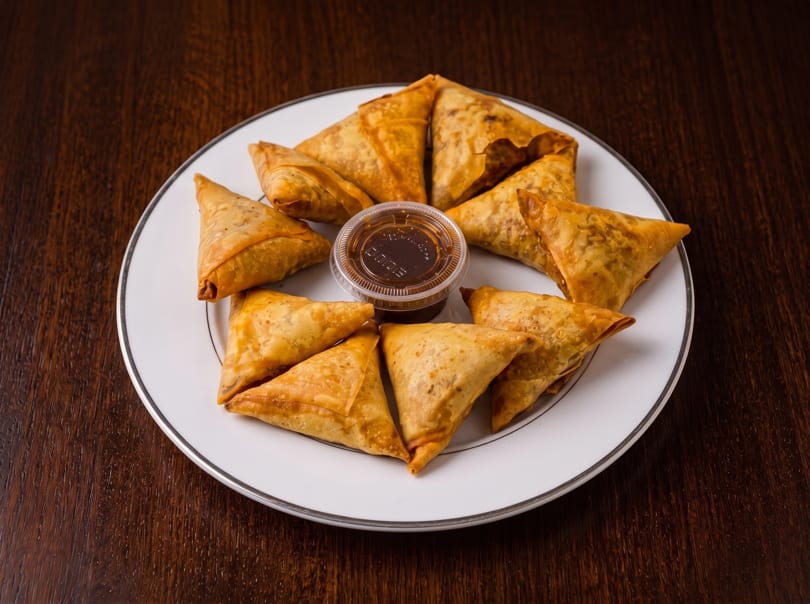 Golden-brown samosas arranged on a white plate with a small container of dipping sauce.