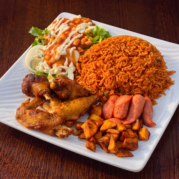 A white rectangular plate with jollof rice, fried chicken, plantains, sausage, and a side salad.
