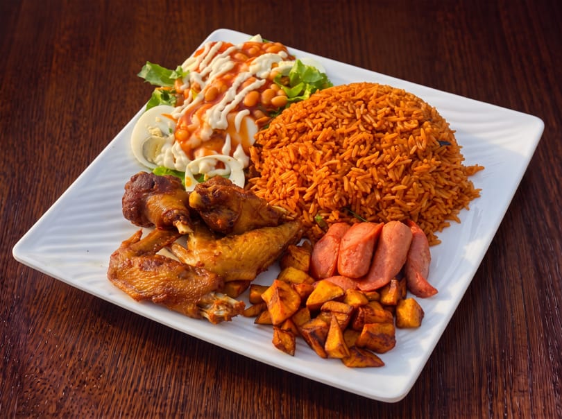 A white rectangular plate with jollof rice, fried chicken, plantains, sausage, and a side salad.