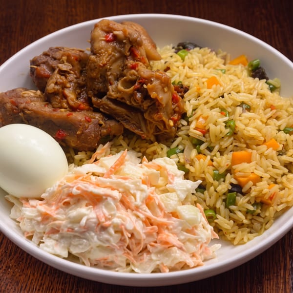 A white bowl with chicken, rice, coleslaw, and a hard-boiled egg on a wooden table.
