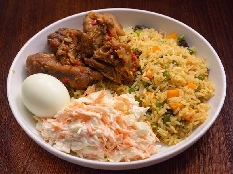 A white bowl with chicken, rice, coleslaw, and a hard-boiled egg on a wooden table.