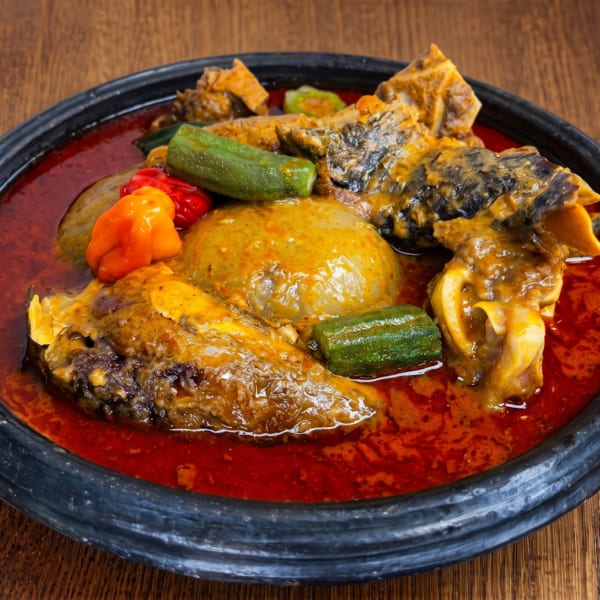 A dark bowl of Ghanaian fufu with red soup, meat, okra, and peppers on a wooden table.
