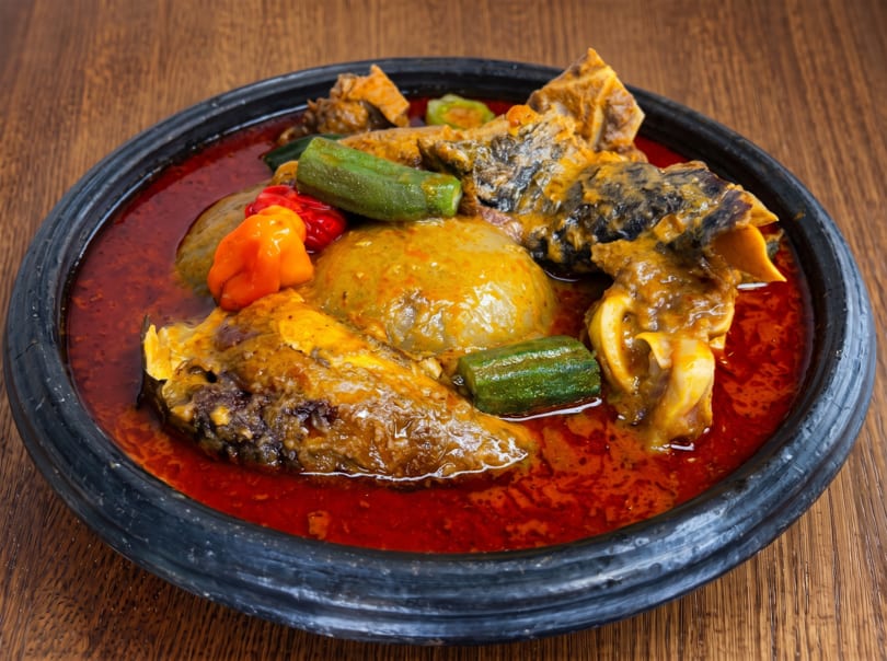 A dark bowl of Ghanaian fufu with red soup, meat, okra, and peppers on a wooden table.