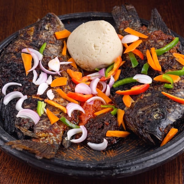 Two whole grilled fish with fufu, vegetables, and sauce on a dark platter.