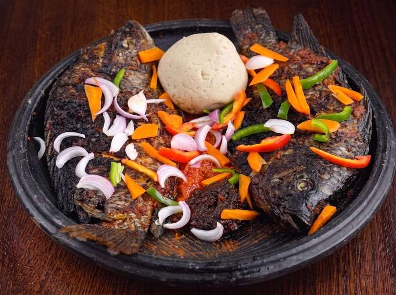 Two whole grilled fish with fufu, vegetables, and sauce on a dark platter.