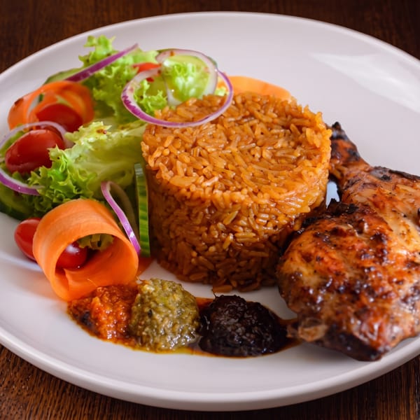 Grilled chicken, jollof rice, fresh salad, and three sauces on a white plate.