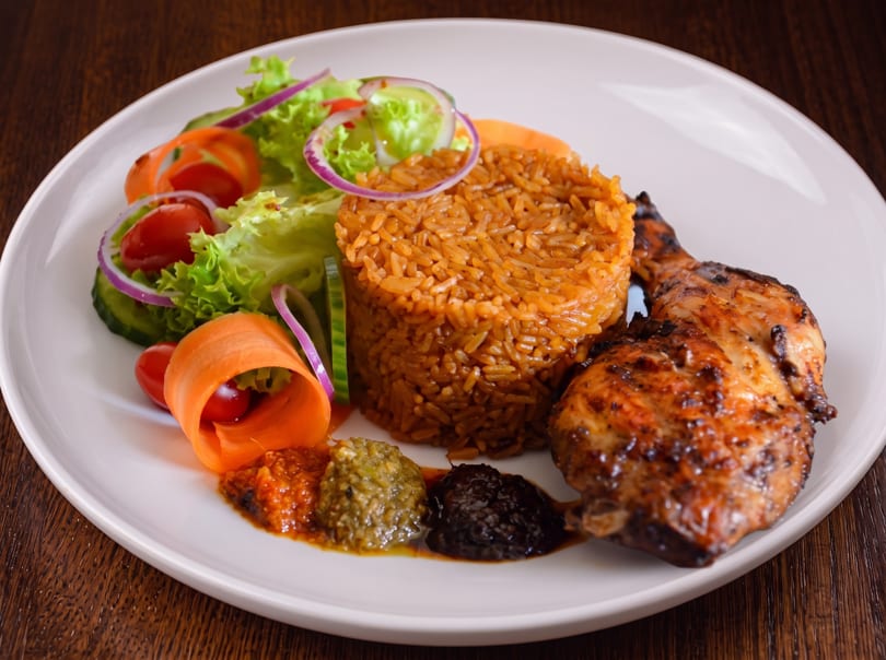 Grilled chicken, jollof rice, fresh salad, and three sauces on a white plate.