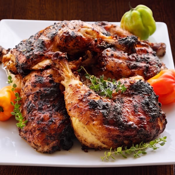 Grilled chicken pieces with charred skin, garnished with fresh herbs and colorful habanero peppers on a white plate.