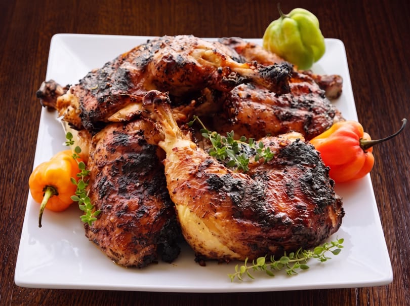 Grilled chicken pieces with charred skin, garnished with fresh herbs and colorful habanero peppers on a white plate.