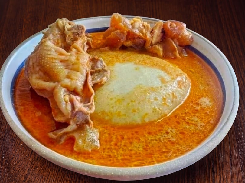 A plate of fufu and chicken in a rich orange soup on a wooden table.
