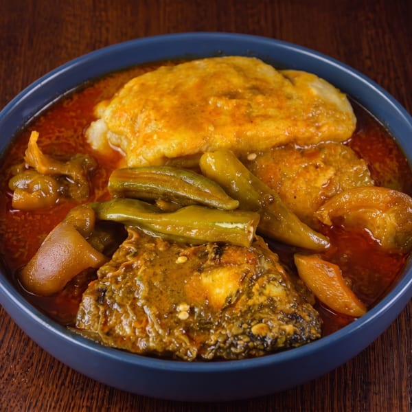 A blue bowl filled with a rich red stew, okra, fish, and fufu on a wooden table.