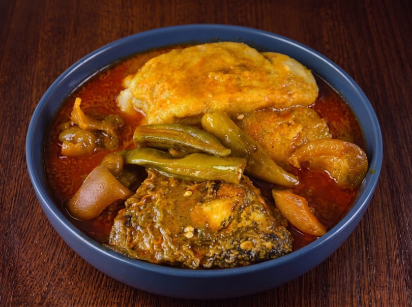 A blue bowl filled with a rich red stew, okra, fish, and fufu on a wooden table.